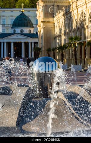 Marianske Lazne, Repubblica Ceca - Maggio 30 2021: Vista della fontana d'acqua che spurta, edificio giallo colonnato e Cross Spring Pavilion sullo sfondo. Foto Stock