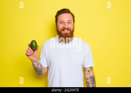 L'uomo felice con barba e tatuaggi tiene un avocado Foto Stock