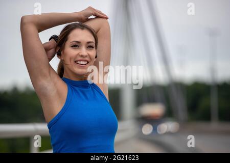 Giovane donna sorridente che si esercita fuori sul ponte. Atleta flessibile nel fuoco che tiene il gomito del braccio di stiramento. Foto Stock