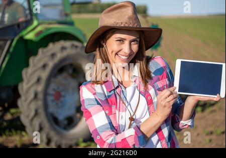 Giovane donna che usa la tecnologia moderna in agricoltura guardando la macchina fotografica. Coltivatore in piedi sul campo davanti al trattore con il tablet. Foto Stock