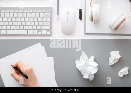 La mano scrive note su un foglio di carta nell'area di lavoro moderna. Frammenti di carta dopo la scrittura sulla scrivania da ufficio con tastiera e mouse con cuffie Foto Stock