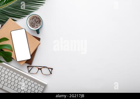 Schermo mock-up smartphone, nota tastiera bicchieri da caffè bicchieri verde pianta foglie di palma sulla scrivania bianca spazio di lavoro. Telefono cellulare sulla moderna scrivania dell'ufficio Foto Stock