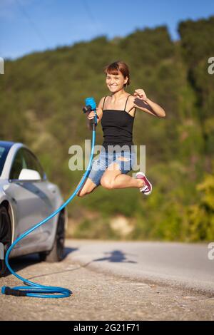 Bella giovane ragazza che salta accanto a un'auto elettrica. Tenendo il cavo di ricarica blu. Foto Stock