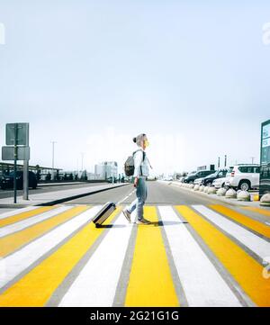 Giovane donna che indossa jeans che attraversano la strada da sola con maschera in cotone giallo e valigia grigia da viaggio in mano. Concetto di turismo del passaporto di Covid Foto Stock