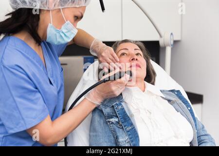 Una giovane cosmetologa femminile in una maschera protettiva fa di una donna anziana cliente una pulizia sottovuoto del viso Foto Stock