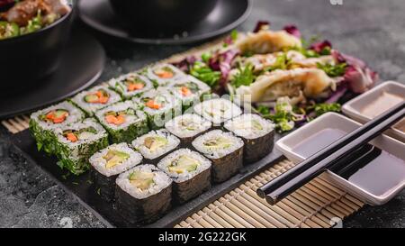 Banner alimentare: Gustoso cibo tradizionale asiatico. Involtini di sushi maki di verdure, gnocchi di gyoza con insalata su una tavola di pietra nera. Stuoia di bambù, noce e salsa di soia Foto Stock