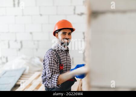 Un lavoratore in cappuccio, tenendo un pettine puty-coltello e coprendo una superficie di parete con pasta adesiva prima di posare pietra decorativa. Lavori di facciata, ristrutturazione e Foto Stock
