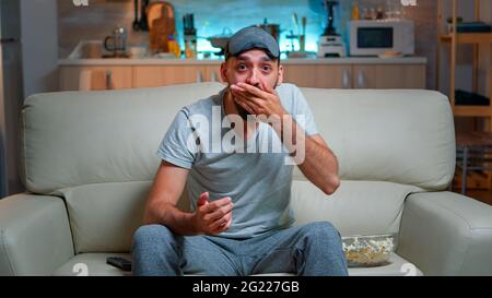 Uomo concentrato con maschera di sonno oculare seduto di fronte alla televisione mentre si guarda film di intrattenimento. Maschio caucasico con barba sdraiata sul divano fino a tarda notte in cucina Foto Stock