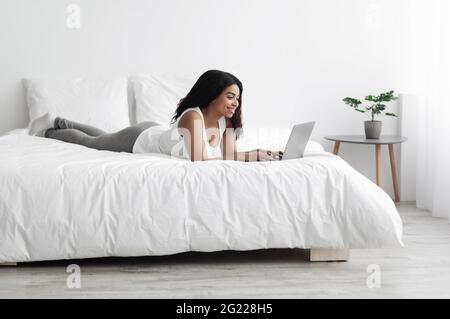 Donna nera distesa a letto e che utilizza un computer portatile, lavora in remoto, chattando o navigando sui social media Foto Stock