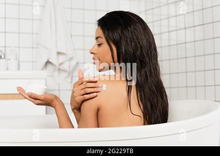 vista laterale della giovane donna afroamericana che tiene il vaso e applica crema sul retro in bagno Foto Stock