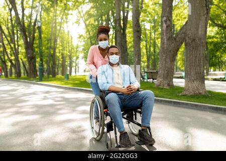 Una donna nera millenaria a piedi con il marito in sedia a rotelle, indossando maschere al parco urbano Foto Stock