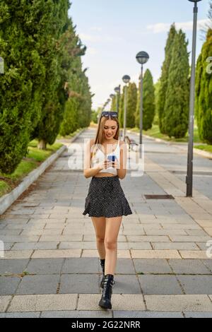 Bella ragazza in abiti estivi a piedi in un parco e chiacchierare sul suo cellulare. Navigare in Internet, visualizzare i social network con il suo smartphone. Concetto di tecnologia. Foto di alta qualità Foto Stock