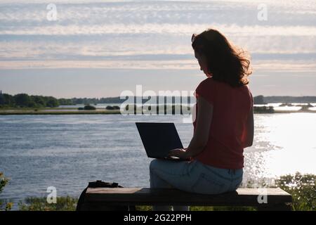 La ragazza si siede su una panchina e utilizza un computer portatile. Comunicazione fuori città. Campo Foto Stock