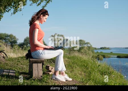 La ragazza si siede su una panchina e utilizza un computer portatile. Comunicazione fuori città. Campo Foto Stock