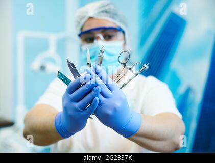 Strumenti dentali in acciaio inossidabile nelle mani di un dentista femminile offuscato che indossa uniforme medica e maschera protettiva e occhiali Foto Stock