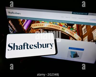 Persona che detiene cellpohone con logo di investimento immobiliare britannico fiducia Shaftesbury plc sullo schermo di fronte alla pagina web. Mettere a fuoco il display del telefono. Foto Stock