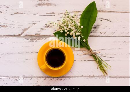 Tavolo da caffè mattutino con bouquet di fiori di Lily of the Valley. Tazza gialla con caffè nero decorato fiori primaverili su tavolo di legno, vista dall'alto Foto Stock