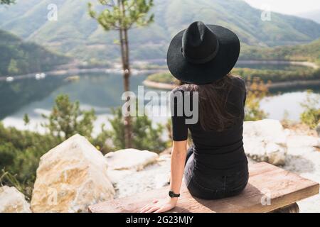 Vista posteriore della donna Traveler seduta su panca di legno e guardando lago di montagna Foto Stock