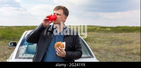 Un uomo alto mangia il pranzo, le bevande da una tazza di plastica nell'aria fresca vicino all'automobile. Foto Stock