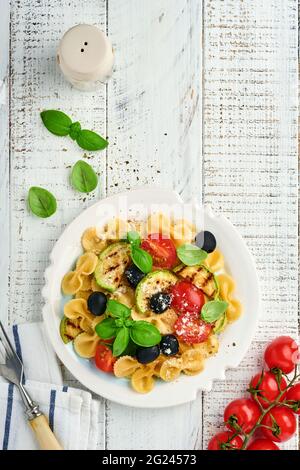 Insalata di pasta fiocchi con verdure zucchine grigliate, pomodori ciliegini, olive, basilico e parmigiano in piatto bianco su ardesia leggera, pietra o calcestruzzo Foto Stock