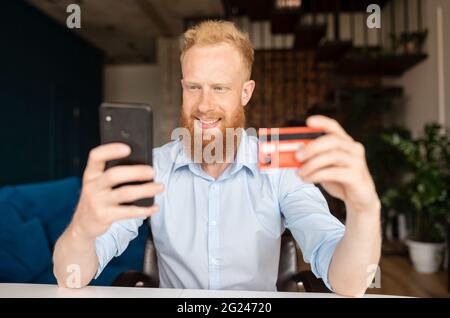 Sereno sorridente rosso testa alato uomo in camicia casual intelligente tenendo carta di credito e smartphone, effettuare transazioni online o il trasferimento di denaro, lo shopping, fare ordine o acquisto, e-banking concetto Foto Stock