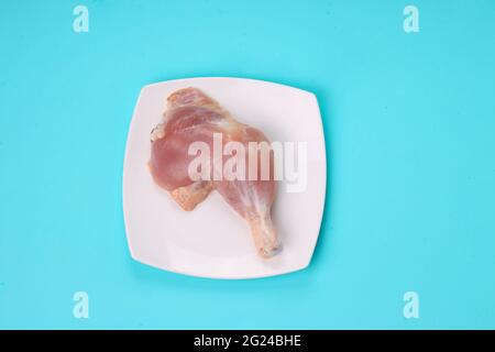 Gamba intera di pollo cruda senza pelle disposta in una tavola bianca di forma quadrata con fondo di colore pieno blu ghiaccio. Foto Stock