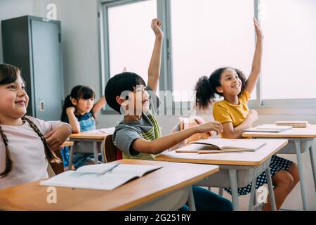 Il ragazzo asiatico di età elementare ha alzato le mani in su in classe. Gruppo diversificato di alunni in età elementare nella scuola di costruzione dell'istruzione. Concetto di volontariato e partecipazione in classe. Foto Stock