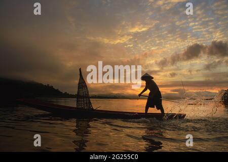 Silhouette pescatore in piedi in una barca gettando rete di pesca nel fiume al tramonto, Thailandia Foto Stock