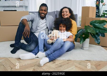 Buona famiglia mista che si diverte mentre si sposta in un nuovo appartamento. Madre, padre e figlia insieme seduti sul pavimento di una nuova casa, con il cardb Foto Stock
