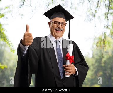 Uomo maturo in una abito di laurea che tiene un diploma e mostra i pollici in su all'aperto Foto Stock