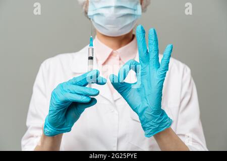 Medico donna o scienziato in bianco camice blu, guanti, cappuccio verde e la maschera contiene una siringa in mani su sfondo bianco. Ella è pronta a dare Foto Stock