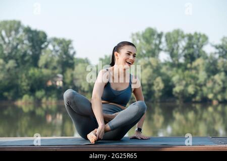 Ridendo giovane donna che fa yoga all'aperto in un bel posto su un lato del fiume. Alberi che si riflettono nell'acqua. In una giornata di sole sotto un blu chiaro Foto Stock