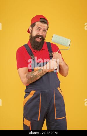 felice uomo bearded pittore operaio in uniforme di caldaietta usando l'attrezzo del rullo di vernice per verniciare la parete nel colore giallo, decoratore Foto Stock