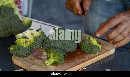 Mano con coltello, taglio broccoli, verdure fresche e crude, verdi e sane su tagliere di legno. Foto Stock