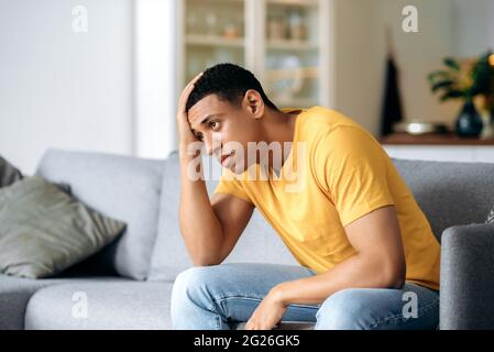 Triste infelice bello ispanico giovane adulto solo uomo, seduto sul divano a casa, tenendo la fronte, con mal di testa, depressione, deluso, ha bisogno di sostegno psicologico Foto Stock