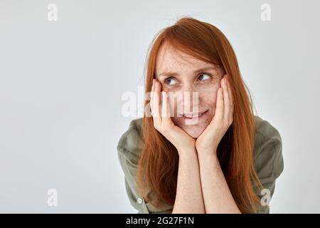 Donna positiva con capelli di zenzero che tiene le mani sulle guance e che guarda via contro sfondo bianco Foto Stock