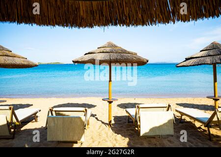 (Fuoco selettivo) splendida vista di alcuni ombrelli di paglia e sedie a sdraio su una spiaggia bagnata da un bel mare turchese. Sardegna, Italia. Foto Stock