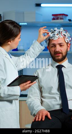 Giovane uomo che indossa cuffie per la scansione delle onde cerebrali che discute con il ricercatore neurologico dei sintomi della malattia. Medico scienziato che scrive le informazioni della scansione del cervello su tavoletta che lavora in laboratorio Foto Stock