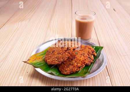 Parippu vada o Lentil Fritters, disposti tradizionalmente in una piastra di acciaio rivestita con foglia di banana e un bicchiere di tè accanto ad esso con legno testurizzato bac Foto Stock