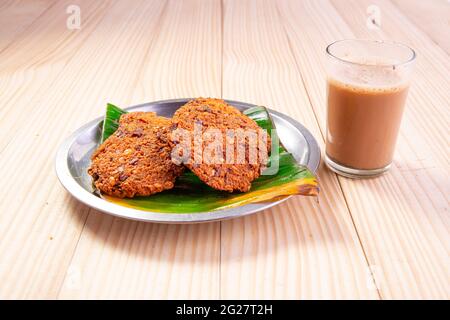 Parippu vada o Lentil Fritters, disposti tradizionalmente in una piastra di acciaio rivestita con foglia di banana e un bicchiere di tè accanto ad esso con legno testurizzato bac Foto Stock