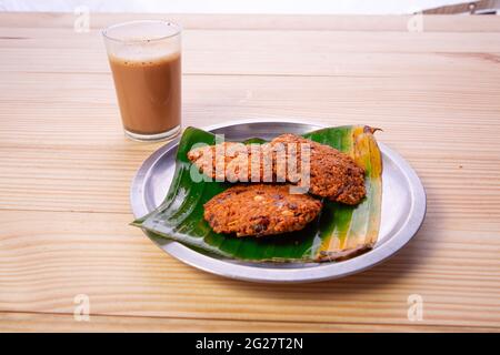 Parippu vada o Lentil Fritters, disposti tradizionalmente in una piastra di acciaio rivestita con foglia di banana e un bicchiere di tè accanto ad esso con legno testurizzato bac Foto Stock