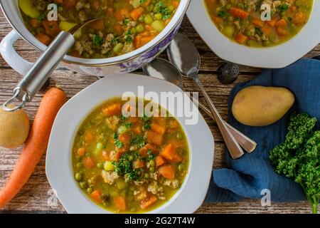 Stufato saporito con piselli e carote, patate e carne tritata servito in una pentola con piatti e cucchiai su sfondo rustico e in legno. Vista dall'alto della tabella Foto Stock