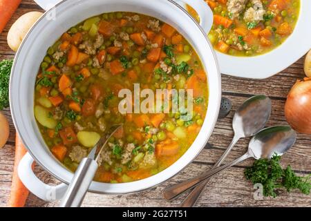 Stufato saporito con piselli e carote, patate e carne tritata servito in una pentola con piatti e cucchiai su sfondo rustico e in legno. Vista dall'alto della tabella Foto Stock