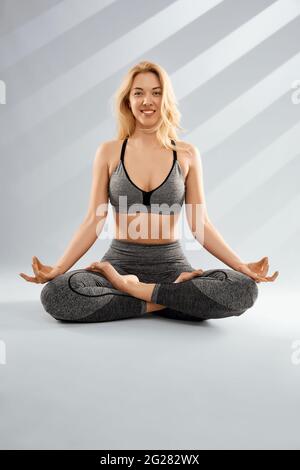 Vista frontale di una giovane bella donna bionda che pratica yoga e meditazione in abbigliamento sportivo alla moda su sfondo grigio. Concetto di stretching e sport per migliorare il benessere. Foto Stock