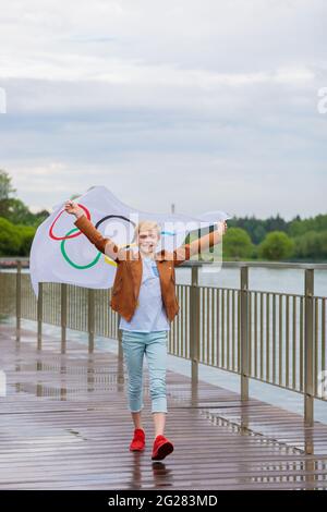 La bionda che sventolava la bandiera olimpica all'aperto nel parco. Giochi Olimpici estivi. Mosca Russia 28 maggio 2021 Foto Stock