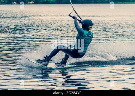 Ragazza con casco e giubbotto di salvataggio fa un wakeboard sul lago con foresta sullo sfondo, wakeboarding, sport acquatici in estate Foto Stock