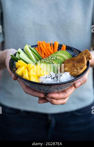 Una femmina in una camicia blu che tiene una ciotola di poke fresca con mango, avocado e tofu, ciotola vegana, verticale Foto Stock