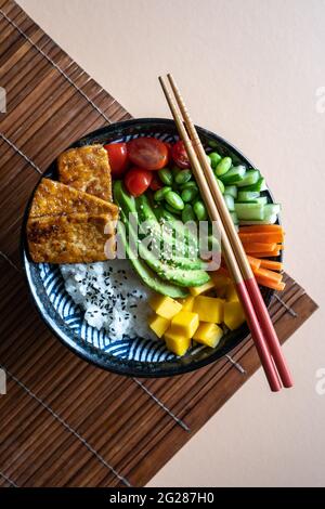 Flatlay di una ciotola di vegan fatta in casa con tofu, mango, avocado, carota, edamame, pomodoro, cetriolo su fondo di mandorla e bambù, verticale Foto Stock