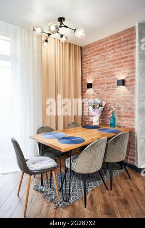 Interni di design per sala da pranzo con sedie in velluto grigio e tavolo in legno, su sfondo di tulle bianco con tenda beige e parete laterale con imitazione file in mattoni. Foto Stock