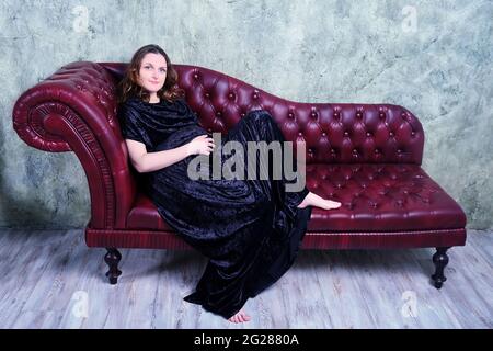 Ritratto di una donna in gravidanza felice seduta su un divano retro in pelle rossa. Donna adulta sorridente in abito da sera lungo e mobili vintage, spazio copia Foto Stock